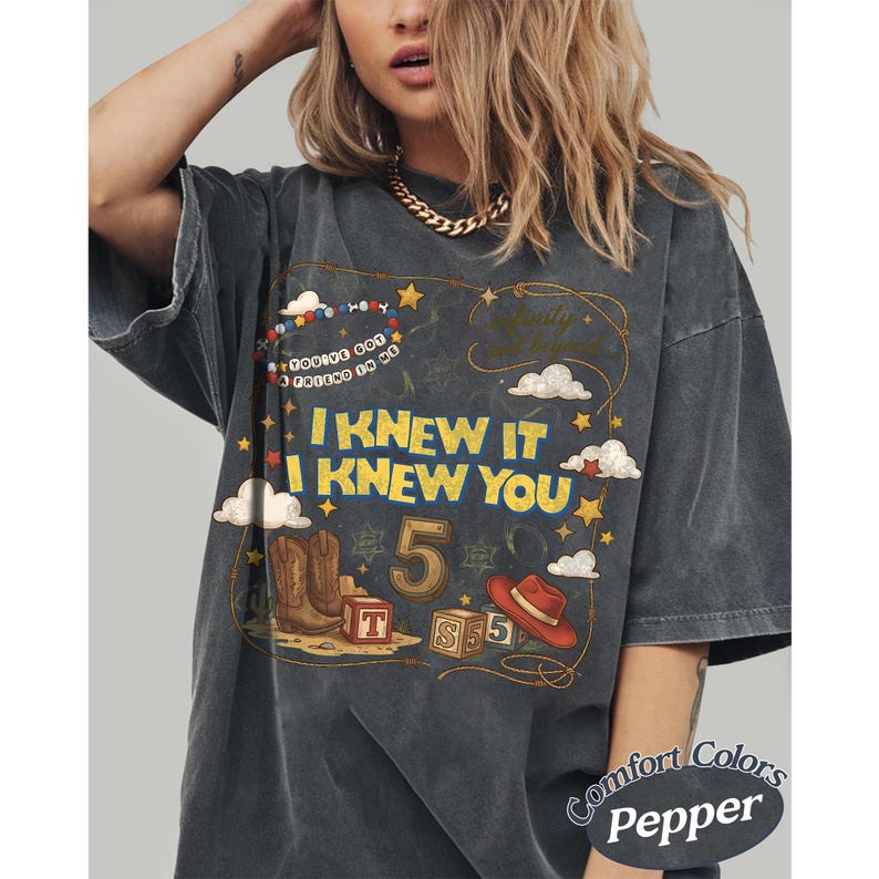 May include: A charcoal gray t-shirt featuring a graphic print with the text I KNEW IT I KNEW YOU, YOU'VE GOT A FRIEND IN ME, and infinity and beyond. The design includes a cowboy boot, a red hat, alphabet blocks, the number 5, and stars.