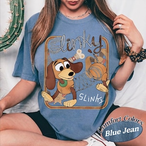 May include: A blue t-shirt featuring a graphic of a brown dachshund toy with a metal spring body. The design includes the text Slinky in a stylised font, a bone, and a sheriff star inside a brown rectangular border. The shirt has a crew neckline.
