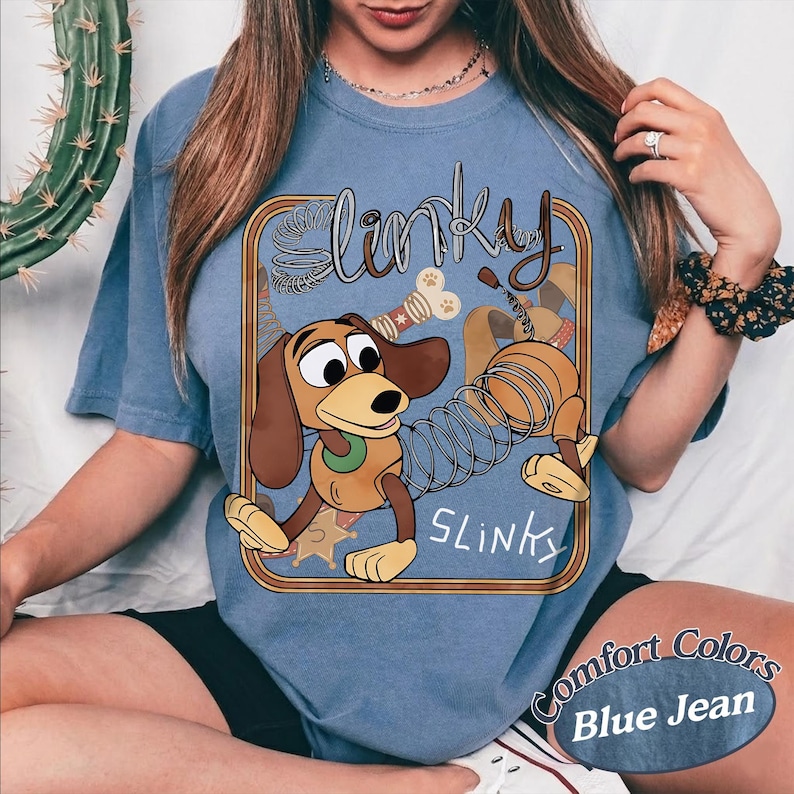 May include: A blue t-shirt featuring a graphic of a brown dachshund toy with a metal spring body. The design includes the text Slinky in a stylised font, a bone, and a sheriff star inside a brown rectangular border. The shirt has a crew neckline.