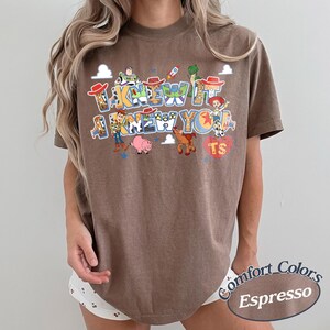 May include: A brown short-sleeve t-shirt featuring a graphic print of various Toy Story characters including Woody, Buzz Lightyear, Jessie, Rex, Bullseye, and Hamm. The text reads I KNEW IT I KNEW YOU with a red heart containing the letters TS.