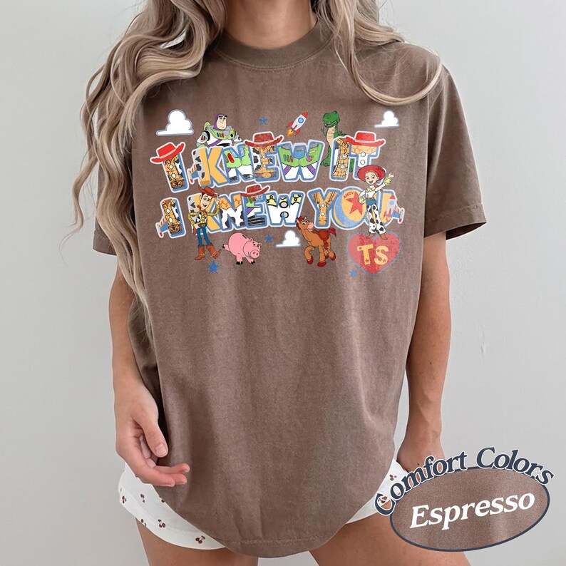 May include: A brown short-sleeve t-shirt featuring a graphic print of various Toy Story characters including Woody, Buzz Lightyear, Jessie, Rex, Bullseye, and Hamm. The text reads I KNEW IT I KNEW YOU with a red heart containing the letters TS.
