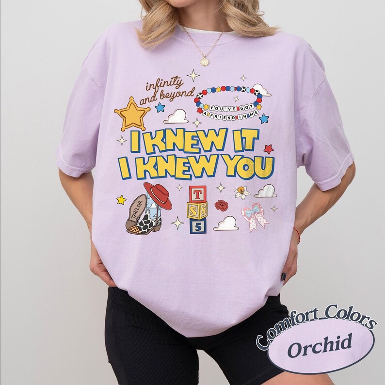 May include: An orchid-coloured t-shirt featuring a graphic print with the text "infinity and beyond," "YOU'VE GOT A FRIEND IN ME," "I KNEW IT," and "I KNEW YOU." The design includes a yellow star, a beaded bracelet, a cowboy boot, alphabet blocks, and a bow.