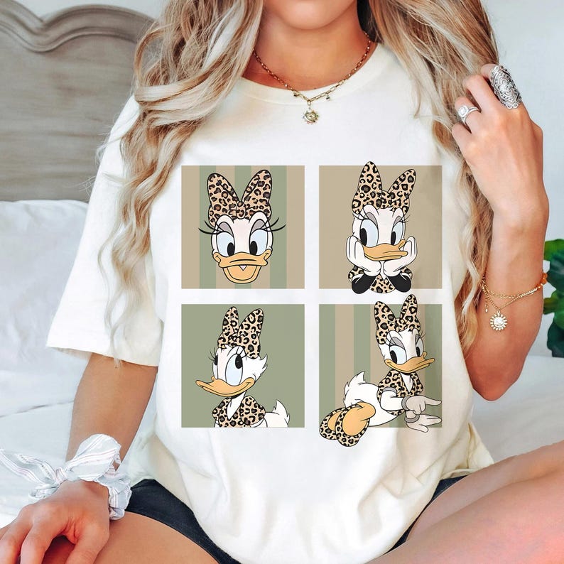 May include: A cream-coloured t-shirt displaying a four-panel design of Daisy Duck in different poses. Each panel shows Daisy with a leopard print bow and accents. The background has vertical stripes in shades of green and beige.