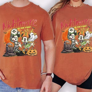 May include: Burnt orange t-shirts featuring a Halloween-themed graphic. The design includes Mickey and Minnie Mouse dressed as skeletons, a pumpkin, and the text "Nightmare on Main Street."