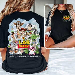 May include: Black t-shirt featuring a colorful graphic of Toy Story characters in lab coats with the words "LAB STORY" and "TO INFINITY AND BEYOND FOR OUR PATIENTS". A smaller graphic on the chest reads "LAB WEEK 2020".