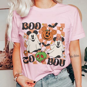 May include: A light pink t-shirt with a Halloween-themed graphic. The design includes the words "BOO TO YOU" in black, along with cartoon ghosts, a pumpkin, and a green skull. The background is a checkered pattern.