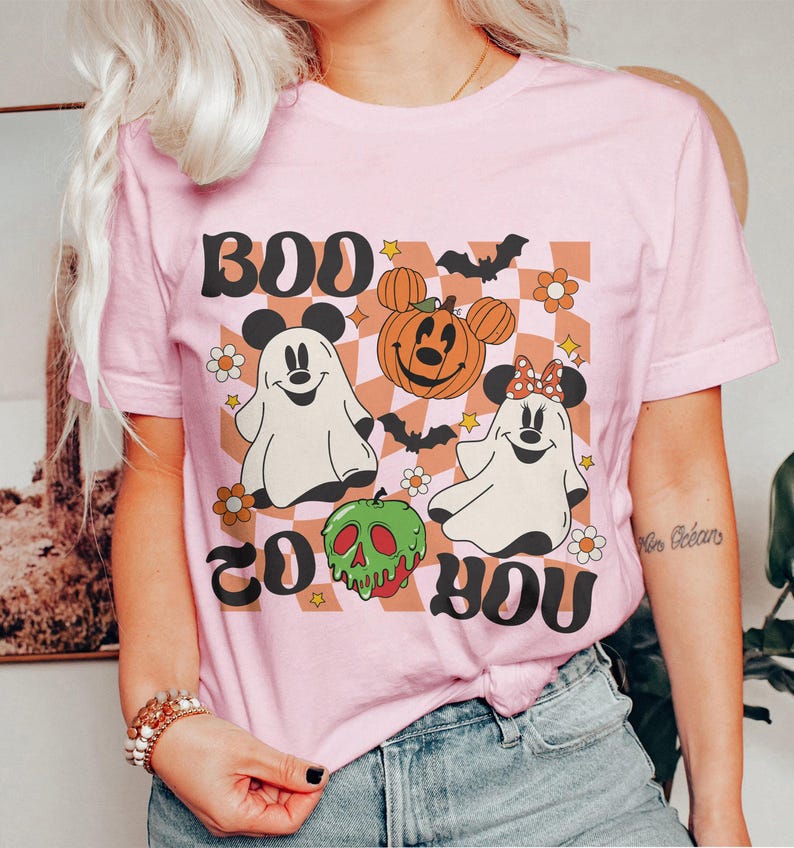 May include: A light pink t-shirt with a Halloween-themed graphic. The design includes the words "BOO TO YOU" in black, along with cartoon ghosts, a pumpkin, and a green skull. The background is a checkered pattern.