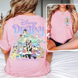 May include: Pink t-shirt featuring the words "Disney Destiny" above a graphic of a cruise ship with Disney characters. The shirt also includes the text "The Smith Family" and the year "2026". A smaller image on the right shows a similar shirt.