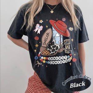 May include: A black short-sleeve t-shirt featuring a graphic print of a cowboy boot with a cow print pattern, a red hat, building blocks with the letters T, S, and the number 13, a pink bow, and a string of beads with the text I KNEW IT I KNEW YOU.
