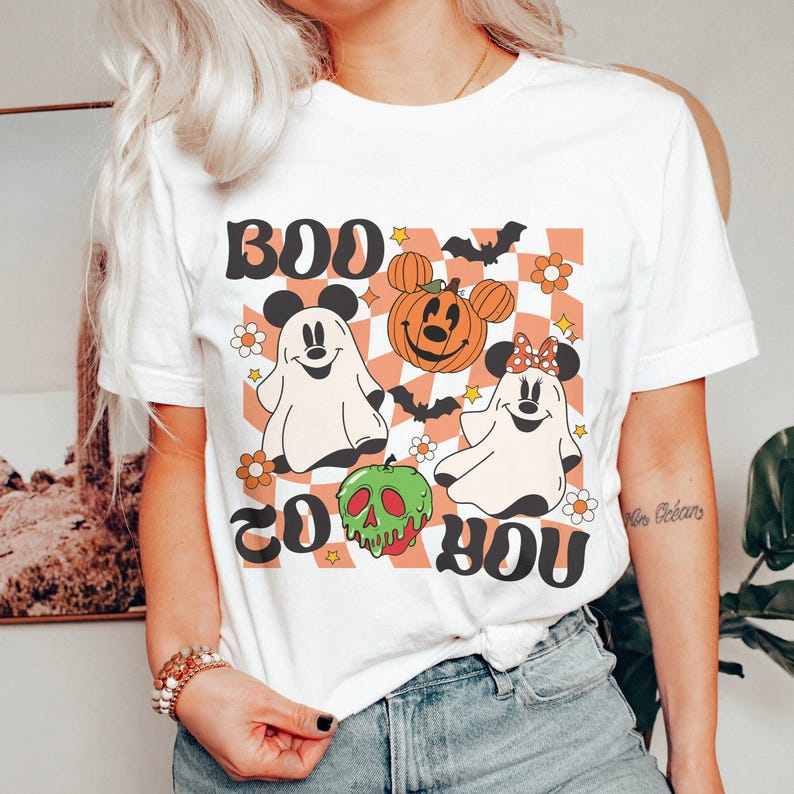 May include: White t-shirt with a Halloween-themed graphic. The design features the words "BOO TO YOU" in a retro font, along with cartoon ghosts, a pumpkin, bats, and flowers. The shirt has a classic crew neck and short sleeves.