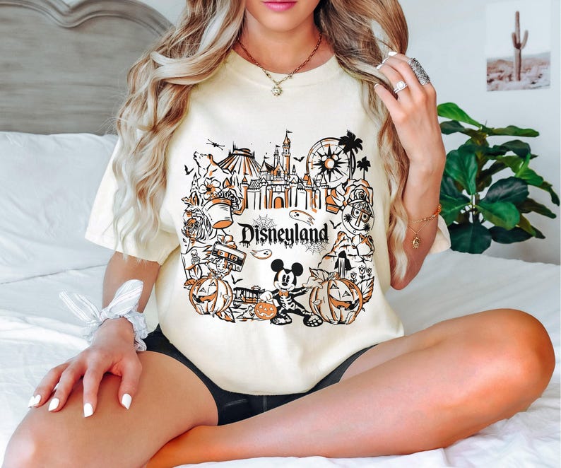 May include: Cream-colored t-shirt featuring a Disneyland graphic in black and orange. The design includes a castle, Mickey Mouse, pumpkins, and the word "Disneyland". The shirt has short sleeves and a relaxed fit.