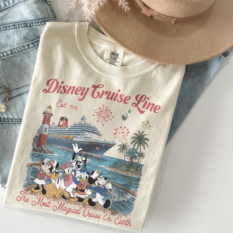 May include: Cream-coloured t-shirt with a Disney Cruise Line graphic. The design features Mickey Mouse, Minnie Mouse, Donald Duck, Daisy Duck, and Goofy, a cruise ship, lighthouse, palm trees, and fireworks. Text reads "Disney Cruise Line Est. 1998" and "The Most Magical Cruise On Earth."