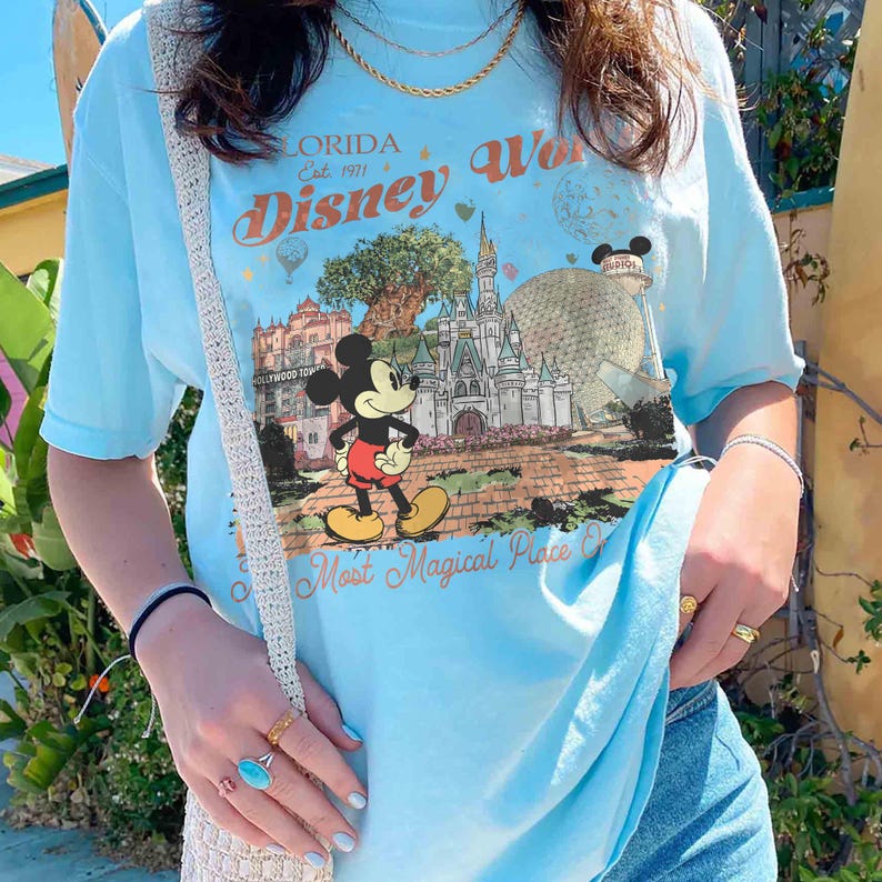 May include: Light blue t-shirt featuring a graphic of Mickey Mouse in front of a castle and other park landmarks. The shirt has the words "Florida Disney World" and "Most Magical Place On Earth" printed on it.