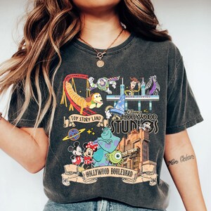 May include: Dark gray t-shirt featuring a colorful graphic of Disney's Hollywood Studios. The design includes characters from Toy Story, Monsters, Inc., and Mickey and Minnie Mouse, with text that reads "Toy Story Land" and "Hollywood Boulevard."