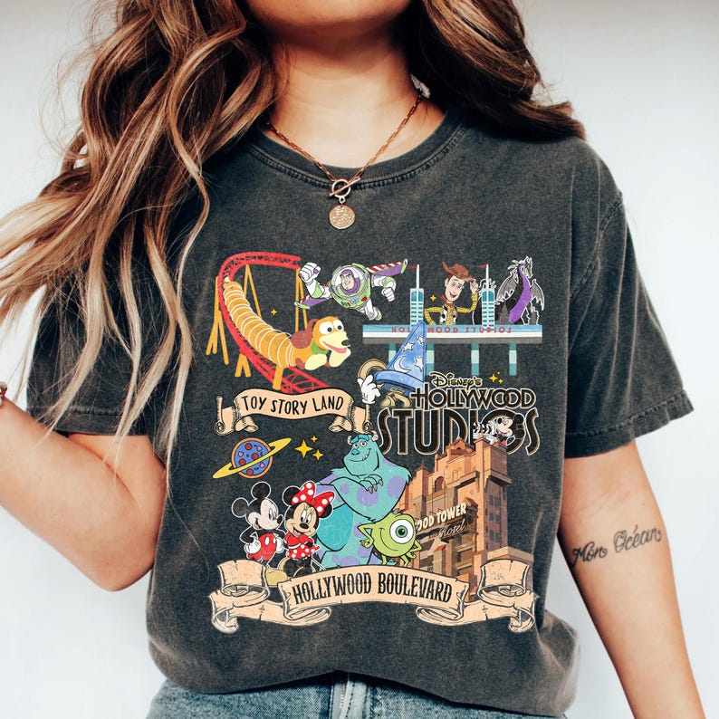 May include: Dark gray t-shirt featuring a colorful graphic of Disney's Hollywood Studios. The design includes characters from Toy Story, Monsters, Inc., and Mickey and Minnie Mouse, with text that reads "Toy Story Land" and "Hollywood Boulevard."