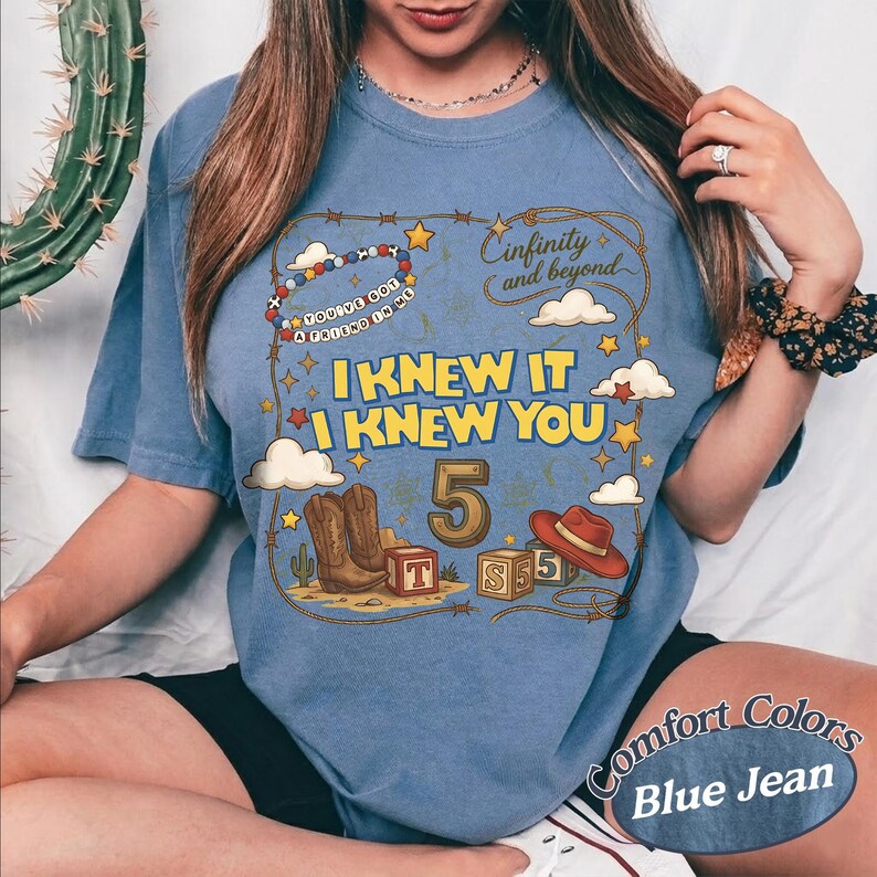 May include: A blue jean colored t-shirt featuring a graphic print with the text "I KNEW IT I KNEW YOU", "5", and "infinity and beyond". The design includes illustrations of cowboy boots, a red hat, alphabet blocks, clouds, and stars.