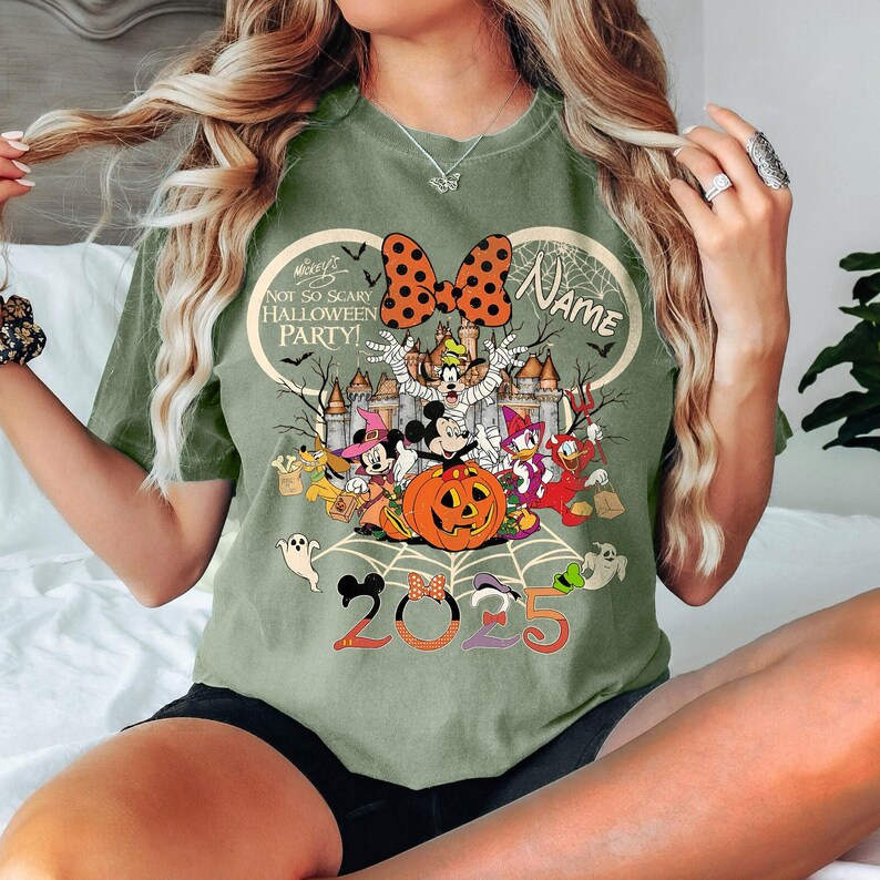 May include: An olive green t-shirt with a Halloween design featuring Disney characters like Mickey and Minnie Mouse. The graphic includes a pumpkin, a castle, and the text "NOT SO SCARY HALLOWEEN PARTY!" and "2025".