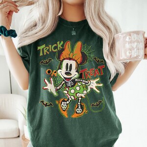 May include: Dark green t-shirt featuring a Halloween-themed Minnie Mouse design. The graphic includes Minnie Mouse in a skeleton costume, the words "Trick or Treat," bats, and a spiderweb. The shirt has a vintage, distressed look.