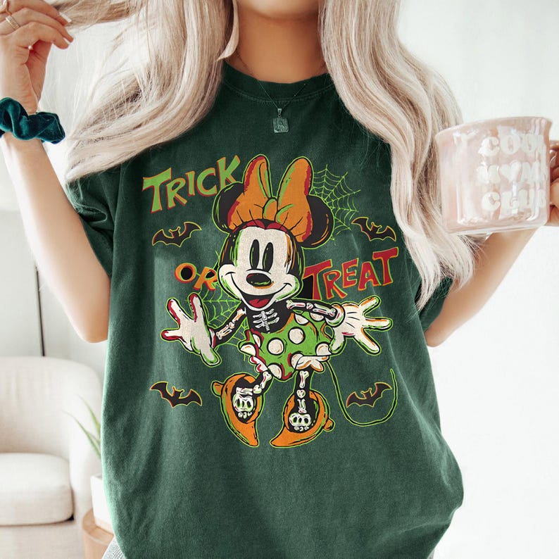 May include: Dark green t-shirt featuring a Halloween-themed Minnie Mouse design. The graphic includes Minnie Mouse in a skeleton costume, the words "Trick or Treat," bats, and a spiderweb. The shirt has a vintage, distressed look.