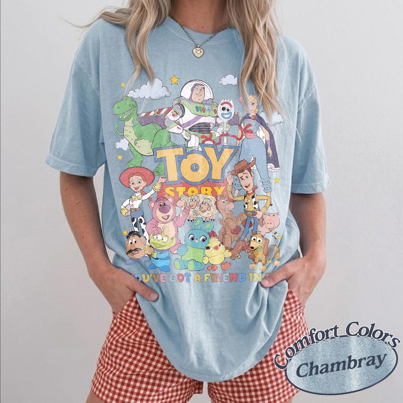 May include: A light blue short-sleeve t-shirt featuring a colorful graphic print of Toy Story characters including Buzz Lightyear, Woody, and Rex. The text reads TOY STORY and YOU'VE GOT A FRIEND IN ME. The shirt has a crew neckline and loose fit.