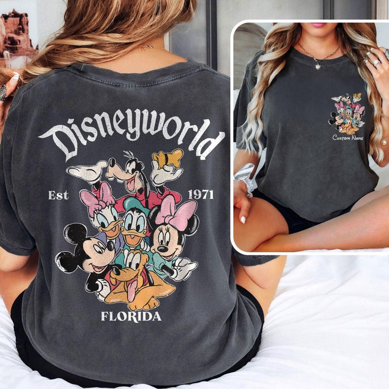May include: Dark gray t-shirt featuring a Disney World design. The back of the shirt displays the words "Disneyworld" above a graphic of Mickey Mouse, Minnie Mouse, Donald Duck, Daisy Duck, Goofy, and Pluto. The text "Est 1971" and "Florida" are also visible.