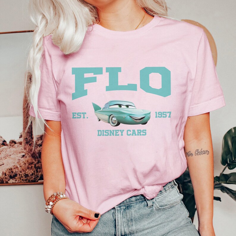 Puede incluir: Camiseta rosa con un gr&aacute;fico azul de un coche turquesa con el texto "FLO EST. 1957 DISNEY CARS".