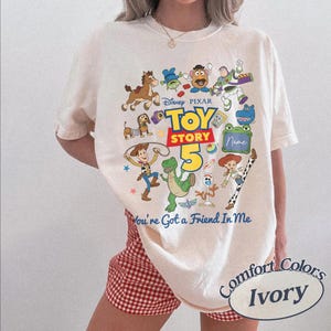 May include: An ivory short-sleeve t-shirt featuring a colorful graphic print of Toy Story characters including Woody, Buzz Lightyear, and Rex. The text reads Disney Pixar Toy Story 5 and You're Got a Friend In Me, with a blank space for a name.