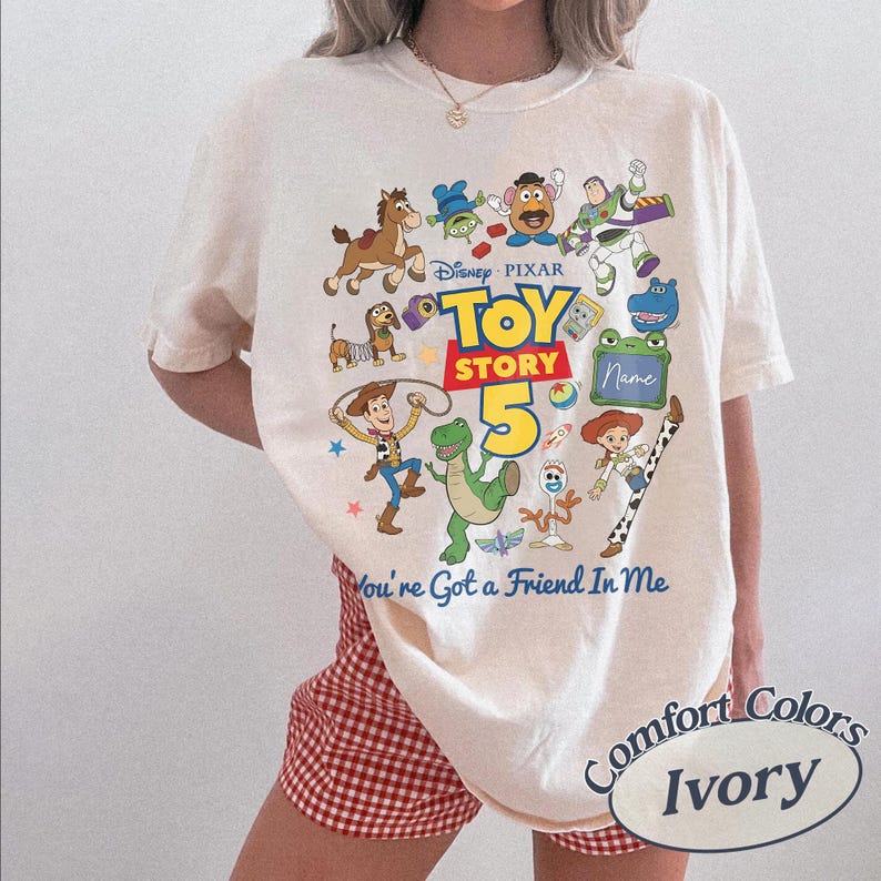 May include: An ivory short-sleeve t-shirt featuring a colorful graphic print of Toy Story characters including Woody, Buzz Lightyear, and Rex. The text reads Disney Pixar Toy Story 5 and You're Got a Friend In Me, with a blank space for a name.