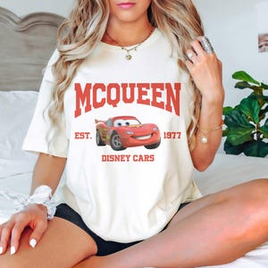 Puede incluir: Camiseta blanca con un gr&aacute;fico rojo de Rayo McQueen de Cars de Disney. El texto "MCQUEEN EST. 1977 DISNEY CARS" est&aacute; impreso en la camiseta.