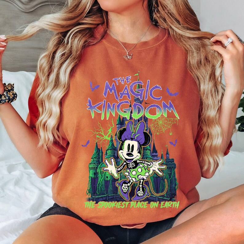 May include: Orange t-shirt featuring a Halloween-themed graphic of Minnie Mouse in front of a castle with the text "The Magic Kingdom" and "The Spookiest Place on Earth". The design includes bats, cobwebs, and green and purple accents.