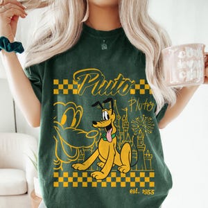 May include: Dark green t-shirt featuring Pluto, the Disney cartoon dog, in yellow. The design includes Pluto's image, the word "Pluto", a castle, fireworks, and the text "est. 1955".