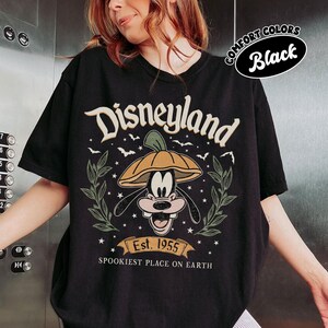 May include: Black t-shirt featuring a Disneyland graphic with Goofy wearing a pumpkin hat. The design includes the text "Disneyland", "Est. 1955", and "Spookiest Place on Earth". The shirt has a round neckline and short sleeves.