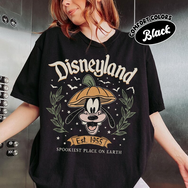 May include: Black t-shirt featuring a Disneyland graphic with Goofy wearing a pumpkin hat. The design includes the text "Disneyland", "Est. 1955", and "Spookiest Place on Earth". The shirt has a round neckline and short sleeves.