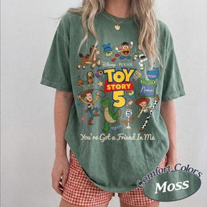 May include: A moss green short-sleeve t-shirt featuring a graphic print of Toy Story characters including Woody, Buzz Lightyear, Rex, and Slinky Dog. The text reads Disney Pixar Toy Story 5 and You're Got a Friend In Me, with a blank name box.