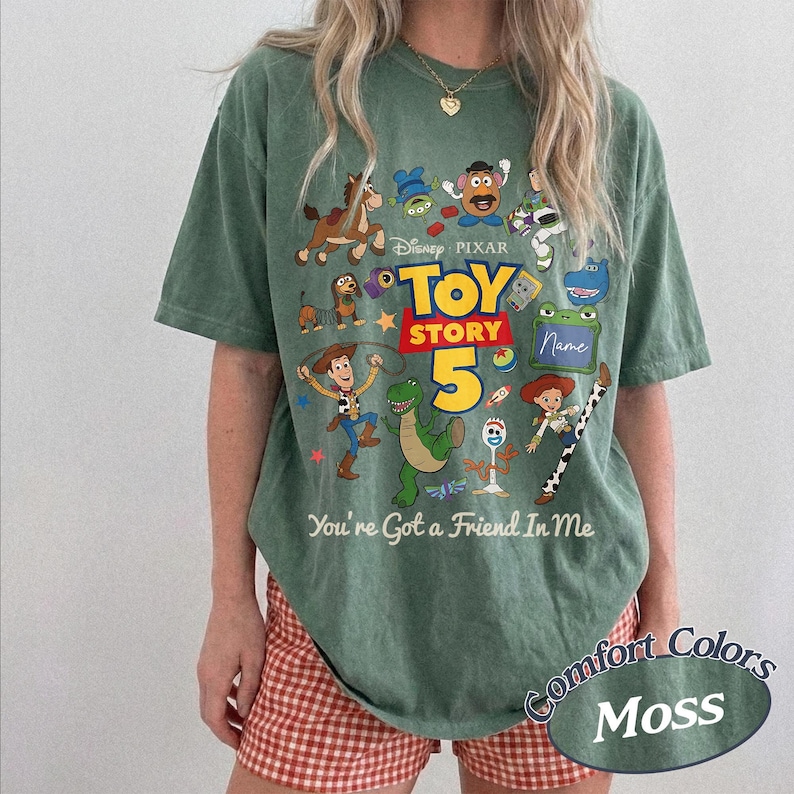 May include: A moss green short-sleeve t-shirt featuring a graphic print of Toy Story characters including Woody, Buzz Lightyear, Rex, and Slinky Dog. The text reads Disney Pixar Toy Story 5 and You're Got a Friend In Me, with a blank name box.
