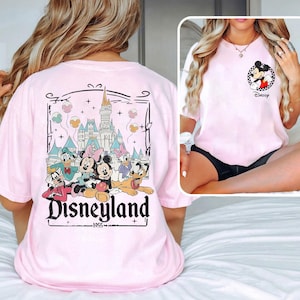 May include: Pink t-shirt with a graphic print of Mickey Mouse and friends in front of Sleeping Beauty's castle. The text "Disneyland 1955" is printed below the graphic.