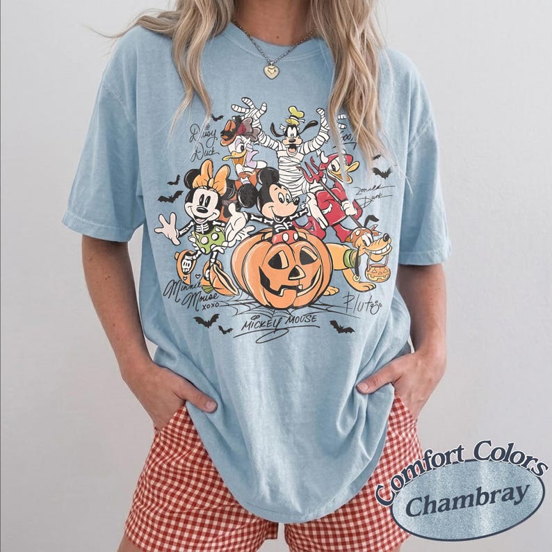 May include: A light blue t-shirt featuring a graphic print of Disney characters in Halloween costumes, including Mickey Mouse sitting on a pumpkin, Minnie Mouse, Goofy, Donald Duck, and Pluto, with bats and spiderwebs, plus the text Comfort Colors Chambray.