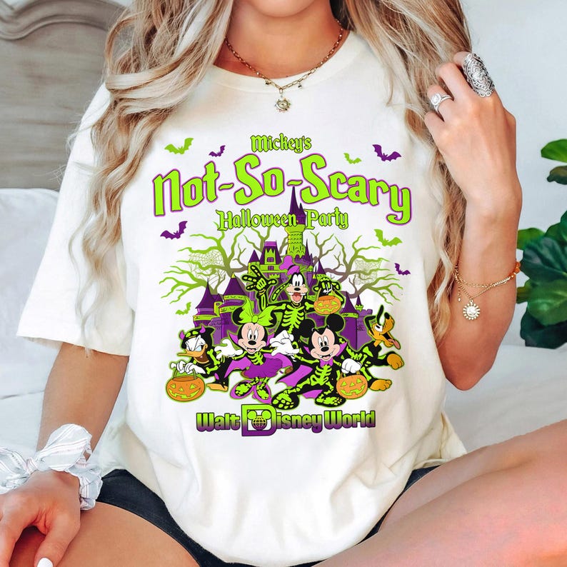 Pu&ograve; includere: Maglietta bianca con una grafica verde e viola di Topolino e i suoi amici vestiti con costumi di Halloween. Il testo sulla maglietta recita "Mickey's Not-So-Scary Halloween Party Walt Disney World".