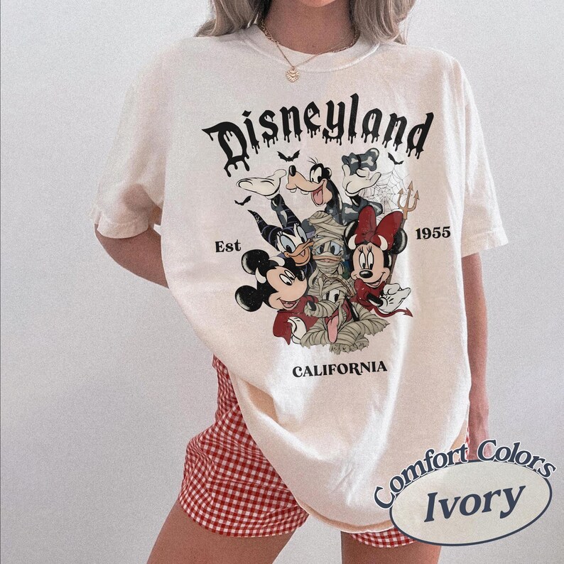 May include: An ivory t-shirt featuring a graphic print of Disney characters in Halloween costumes. The text reads Disneyland, Est 1955, and California. The characters include Mickey Mouse, Minnie Mouse, Donald Duck, Goofy, and a mummy figure.