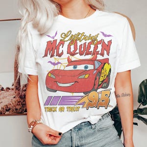 May include: White t-shirt featuring a red cartoon car with the text "Lightning McQueen" and "Trick or Treat". The car has devil horns and the number 95. Purple bats and a spiderweb design are also present.