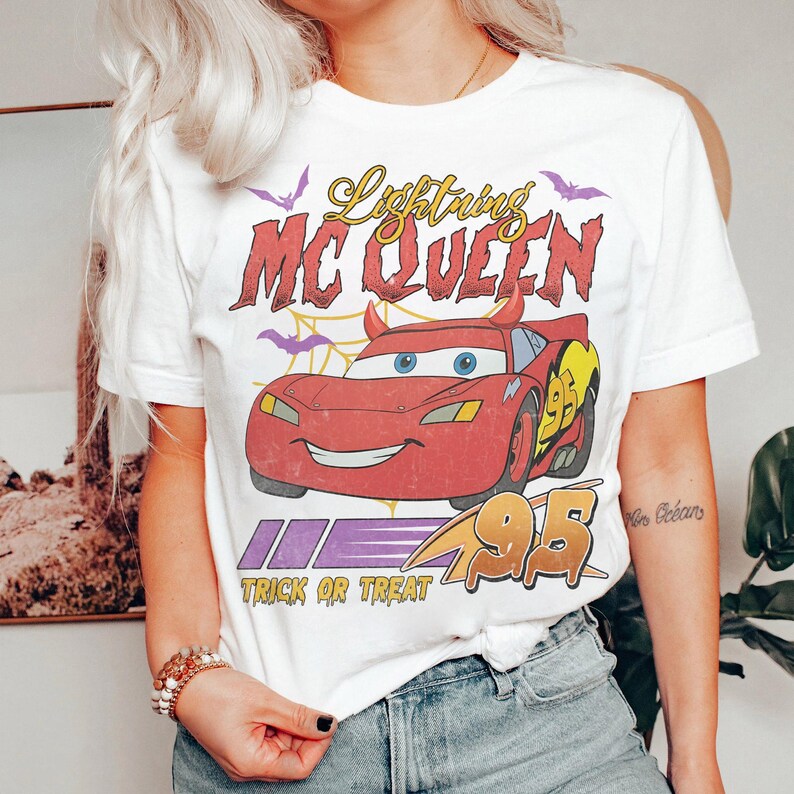 May include: White t-shirt featuring a red cartoon car with the text "Lightning McQueen" and "Trick or Treat". The car has devil horns and the number 95. Purple bats and a spiderweb design are also present.