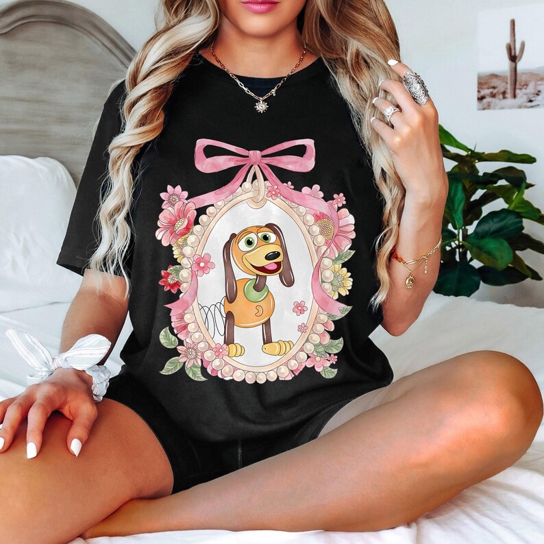 May include: Black t-shirt featuring a cartoon dog character within a pink ribbon-framed oval, surrounded by flowers and pearls. The dog is brown and yellow with a spring body. A cute and colorful design.