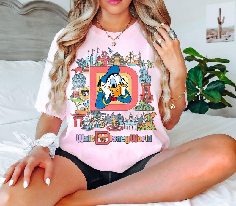 May include: Pink t-shirt featuring Donald Duck inside a large letter "D" with various Disney World landmarks. The words "Walt Disney World" are printed below the graphic in a colorful font. The shirt has short sleeves.