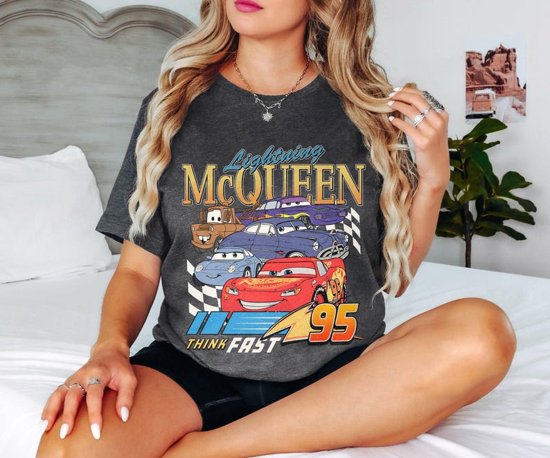 May include: A dark gray t-shirt with a graphic of Lightning McQueen and other Cars characters. The text "Lightning McQueen" and "Think Fast" are printed on the shirt. The number "95" is also printed on the shirt.