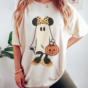 May include: A cream colored t-shirt with a cartoon ghost wearing a yellow bow and carrying a pumpkin. The ghost has black eyes and a black mouth. The pumpkin is orange with a black face.