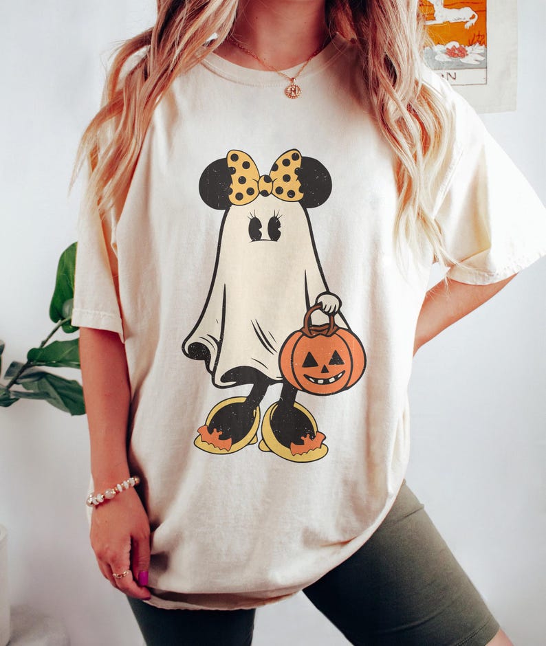 May include: A cream colored t-shirt with a cartoon ghost wearing a yellow bow and carrying a pumpkin. The ghost has black eyes and a black mouth. The pumpkin is orange with a black face.