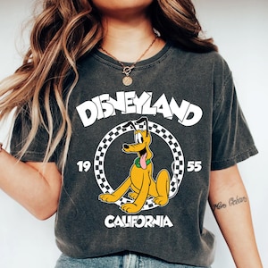 K&ouml;nnte beinhalten: Dunkelgraues T-Shirt mit Pluto von Disney. Das Shirt tr&auml;gt das Wort "Disneyland" in wei&szlig;en Blockbuchstaben &uuml;ber einem kreisf&ouml;rmigen Design mit Pluto und dem Text "1955 California".