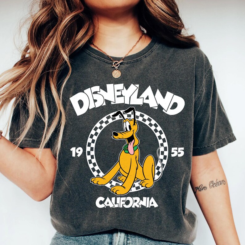 K&ouml;nnte beinhalten: Dunkelgraues T-Shirt mit Pluto von Disney. Das Shirt tr&auml;gt das Wort "Disneyland" in wei&szlig;en Blockbuchstaben &uuml;ber einem kreisf&ouml;rmigen Design mit Pluto und dem Text "1955 California".
