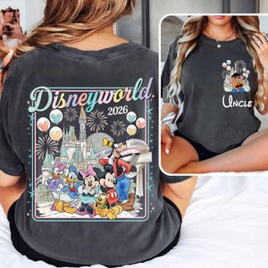 Pu&ograve; includere: T-shirt grigio scuro con una grafica colorata di Disneyworld 2026 sul retro, con fuochi d'artificio, un castello e personaggi dei cartoni animati. La parte anteriore ha una grafica pi&ugrave; piccola di un personaggio con palloncini e la parola "Uncle".