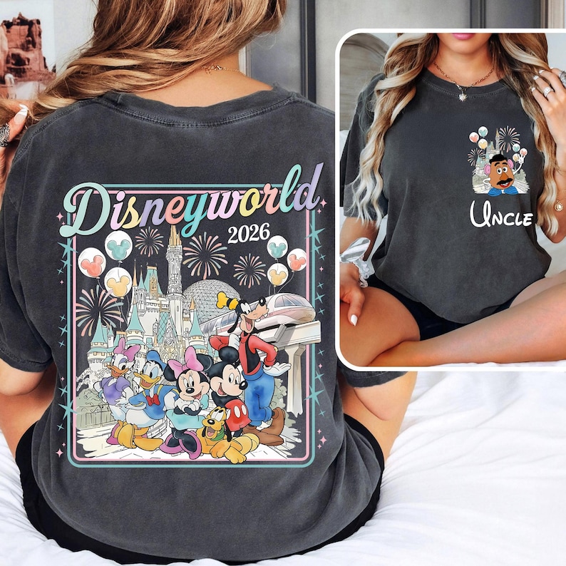 Pu&ograve; includere: T-shirt grigio scuro con una grafica colorata di Disneyworld 2026 sul retro, con fuochi d'artificio, un castello e personaggi dei cartoni animati. La parte anteriore ha una grafica pi&ugrave; piccola di un personaggio con palloncini e la parola "Uncle".